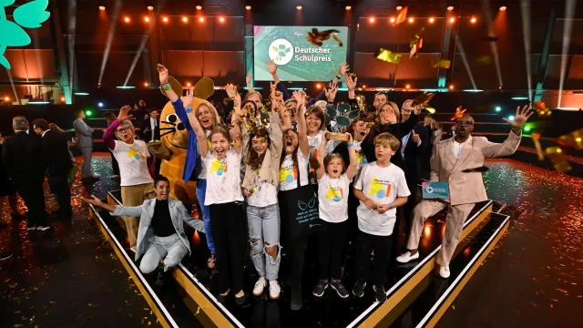 Auf einer Bühne des Deutschen Schulpreises steht eine große Gruppe von Kindern und Erwachsenen. Goldenes Konfetti fällt von der Decke, mehrere Personen halten die Arme in die Höhe. Im Hintergrund sind die Bühne, das Logo des Deutschen Schulpreises und die Figur der Maus zu sehen.