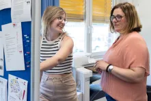Zwei Personen stehen in einem Büro neben einem Drucker und einer Pinnwand mit ausgehängten Dokumenten. Eine Person in einem gestreiften ärmellosen Oberteil zeigt auf die Pinnwand, während die andere Person mit Brille und rosafarbener Bluse danebensteht. Im Hintergrund sind Fenster mit gelben Jalousien zu sehen. Die Szene vermittelt eine Arbeitssituation in einer Schule.