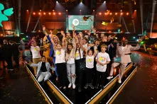 Auf einer Bühne des Deutschen Schulpreises steht eine große Gruppe von Kindern und Erwachsenen. Goldenes Konfetti fällt von der Decke, mehrere Personen halten die Arme in die Höhe. Im Hintergrund sind die Bühne, das Logo des Deutschen Schulpreises und die Figur der Maus zu sehen.