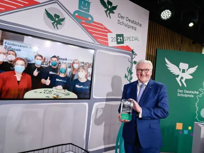 Bei der Verleihung des Deutschen Schulpreises 2021 steht eine Person auf einer Bühne und hält die Preistrophäe in den Händen. Auf einem großen Bildschirm ist das Team der Mosaikschule Marburg per Videoschalte zu sehen. Mehrere Personen tragen Schutzmasken. Im Hintergrund sind das Logo des Deutschen Schulpreises und der Schriftzug „2021 Spezial“ sichtbar.