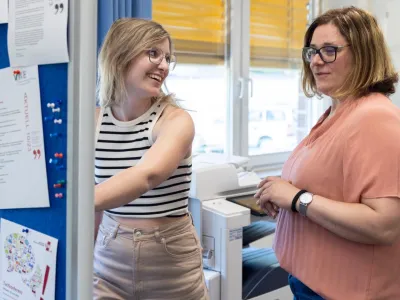 Zwei Personen stehen in einem Büro neben einem Drucker und einer Pinnwand mit ausgehängten Dokumenten. Eine Person in einem gestreiften ärmellosen Oberteil zeigt auf die Pinnwand, während die andere Person mit Brille und rosafarbener Bluse danebensteht. Im Hintergrund sind Fenster mit gelben Jalousien zu sehen. Die Szene vermittelt eine Arbeitssituation in einer Schule.