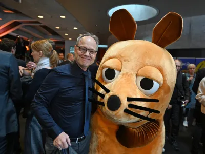 Eine Person in dunklem Hemd steht bei einer Veranstaltung neben der Figur der Maus aus der „Sendung mit der Maus“. Im Hintergrund befinden sich zahlreiche Gäste in einem modern gestalteten Foyer.