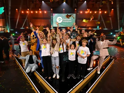 Auf einer Bühne des Deutschen Schulpreises steht eine große Gruppe von Kindern und Erwachsenen. Goldenes Konfetti fällt von der Decke, mehrere Personen halten die Arme in die Höhe. Im Hintergrund sind die Bühne, das Logo des Deutschen Schulpreises und die Figur der Maus zu sehen.