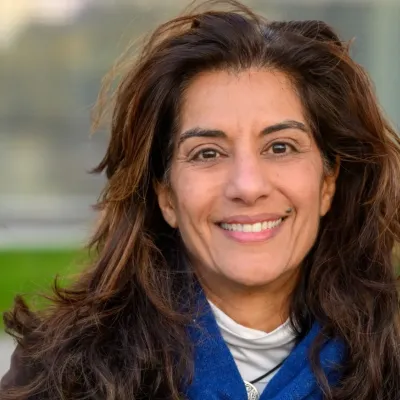 Portrait von Sanam Anderlini: Eine Frau mit langem dunklem Haar vor einem unscharfen Hintergrund im Freien. Sie blickt in die Kamera und trägt einen blauen Schal über einem hellen Oberteil.