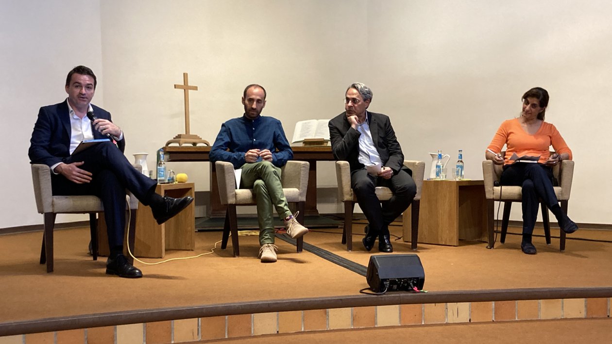 Three men and one woman (right) are sitting in chairs on a podium in a church. The left one speaks to the audience.