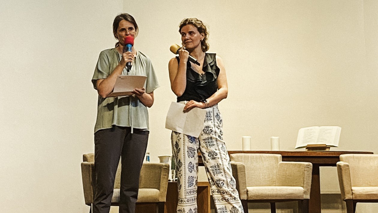 Two women with a microfone, standing side by side, the left one is speaking while the other listens